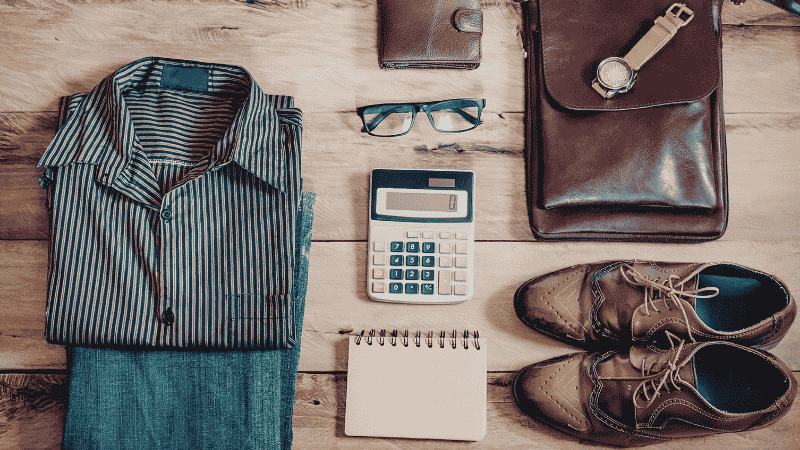 A top-down flat lay of men's wardrobe essentials including a navy hopsack blazer, dark wash indigo denim, a white Oxford button-down shirt, and clean white leather sneakers on a rustic wooden background.