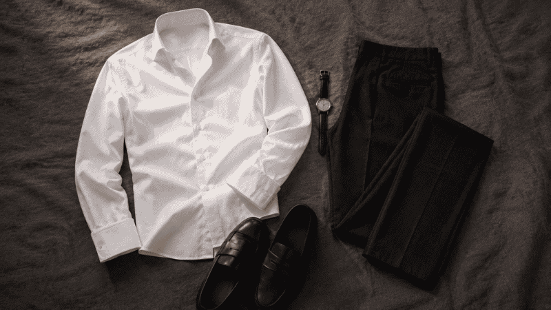 men's first date evening outfit flat lay with white dress shirt open collar dark charcoal trousers and black leather loafers for cocktail bar