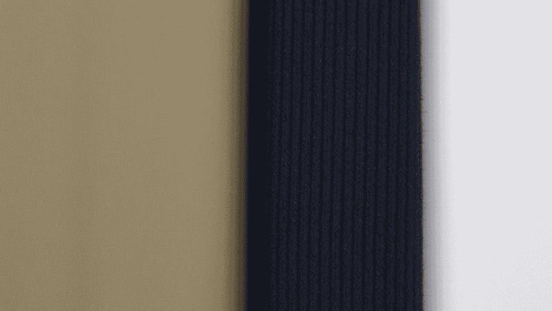 A close-up flat lay of men's style essentials: high-quality khaki cotton twill trousers paired with a navy blue wool sweater and a crisp white Oxford cloth button-down shirt to show color and texture harmony