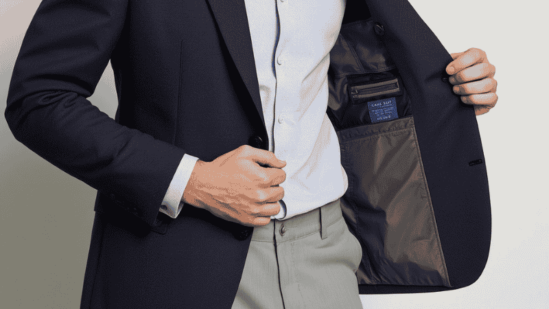 A styling guide photo of a man in a navy blazer and grey trousers, demonstrating the "always top, never bottom" button rule and perfect sleeve length at the wrist bone.