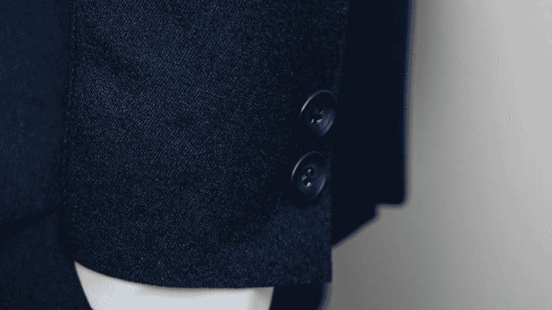 Close-up detail of a navy hopsack wool blazer sleeve, highlighting the matte horn button and the textured weave of the fabric with a white shirt cuff peeking out.